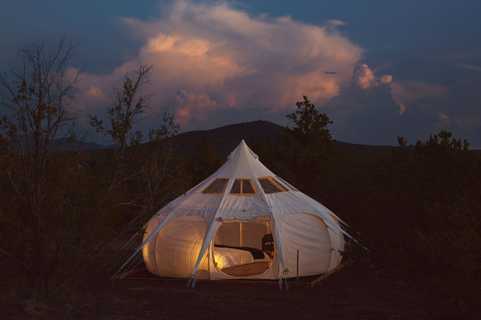 White tent glowing at dusk in a dark field, with mountains and pink clouds in the background