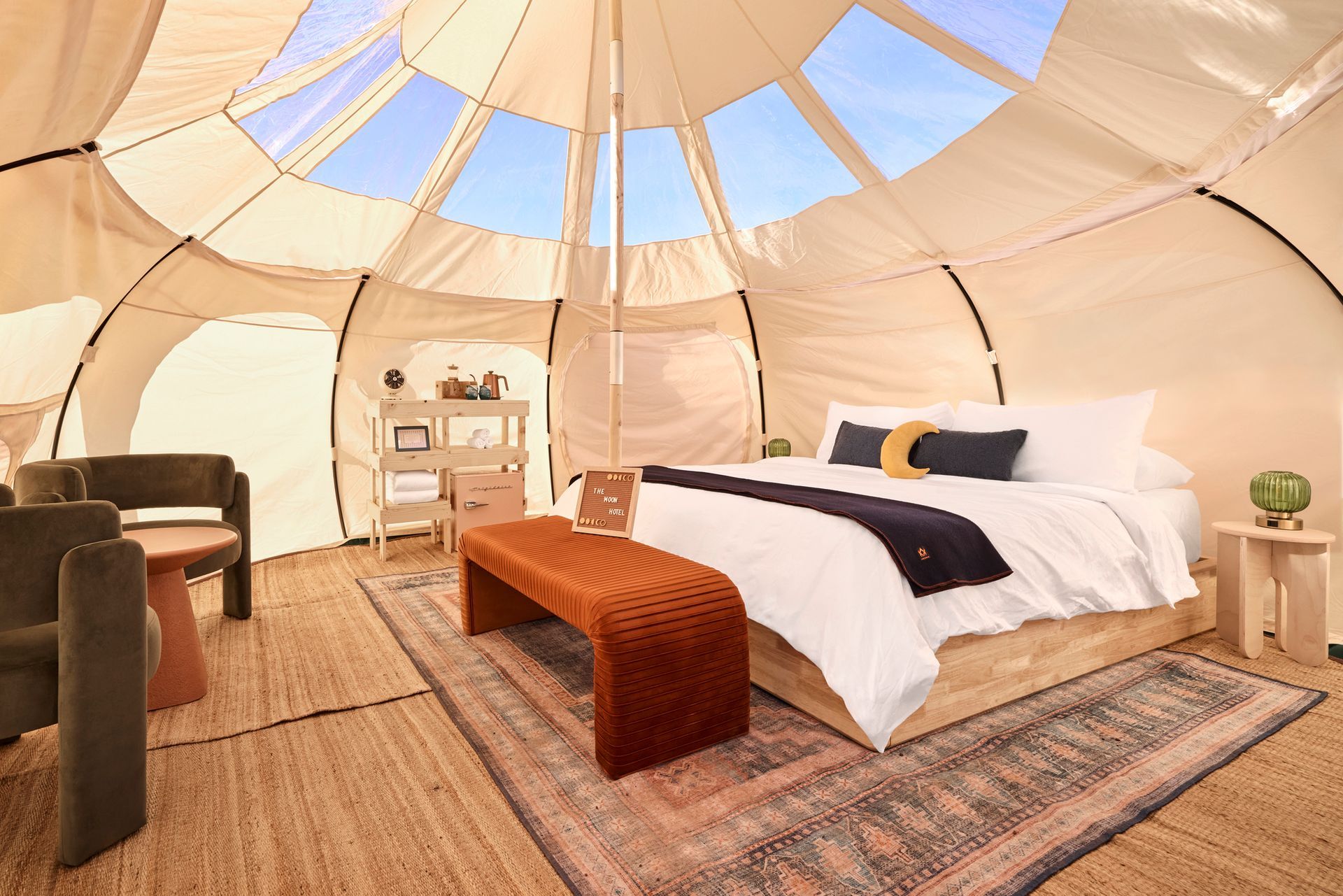 Interior of a glamping tent with bed, bench, rug, two chairs, and a small table. Skylights in ceiling.