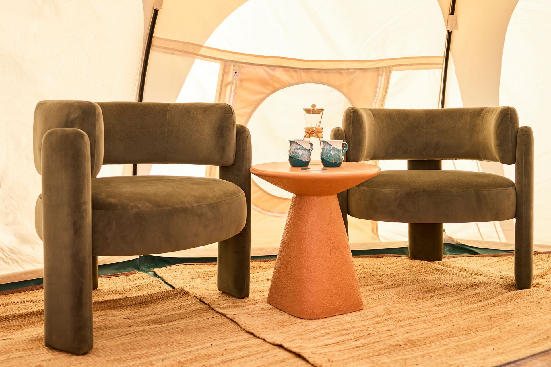 Two olive green armchairs and a small orange table inside a tent.