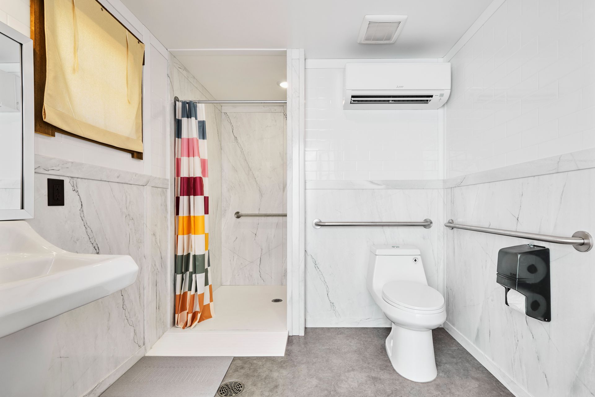 Accessible bathroom with toilet, sink, shower, and grab bars. White and gray tones; shower curtain has colorful stripes.