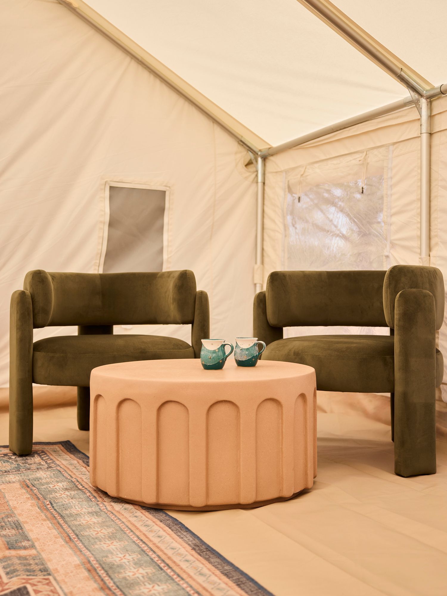 Two green velvet armchairs and a pink coffee table inside a canvas tent.