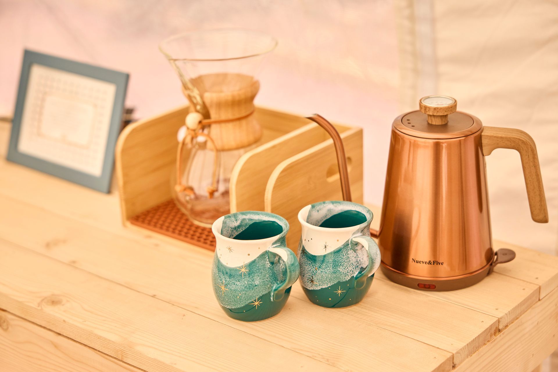 Coffee brewing setup on wood surface: pour-over, copper kettle, turquoise mugs, and a wooden frame.
