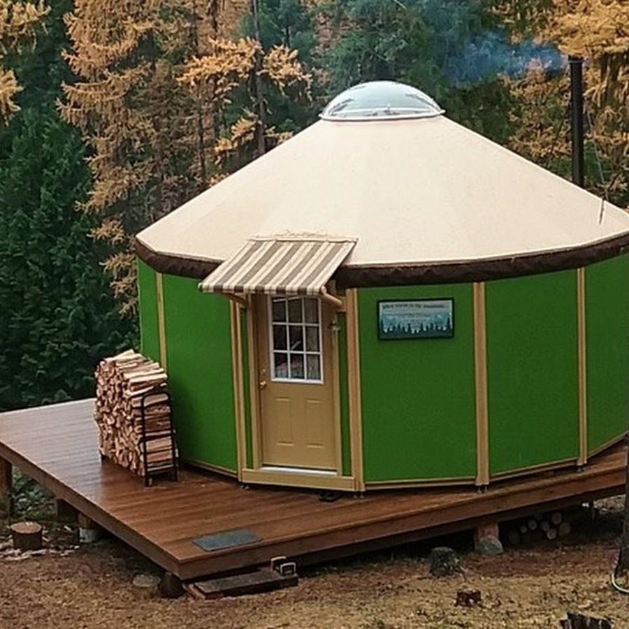 Green yurt with wood deck in a forest. Firewood stacked beside entrance with awning. Dome skylight.
