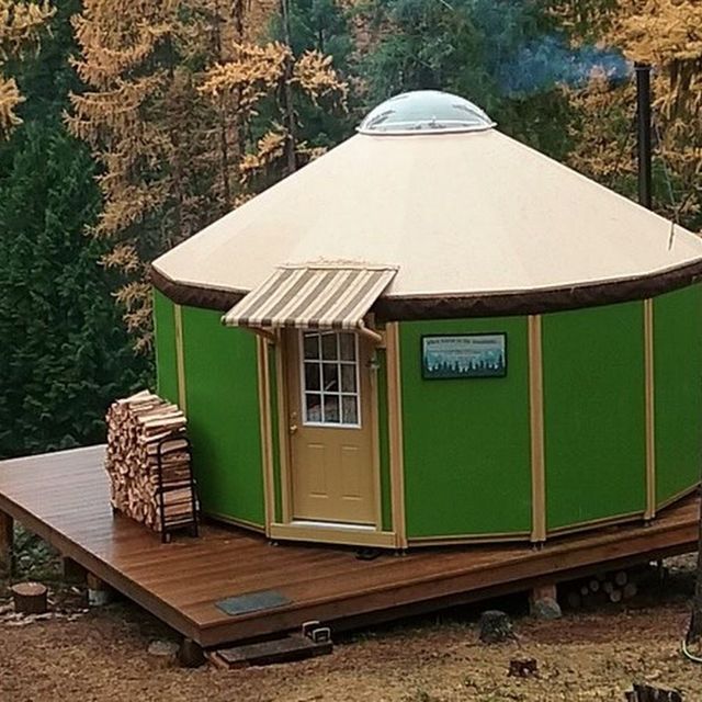 Green round cabin on a wooden deck in the woods, with a tan door and small awning.