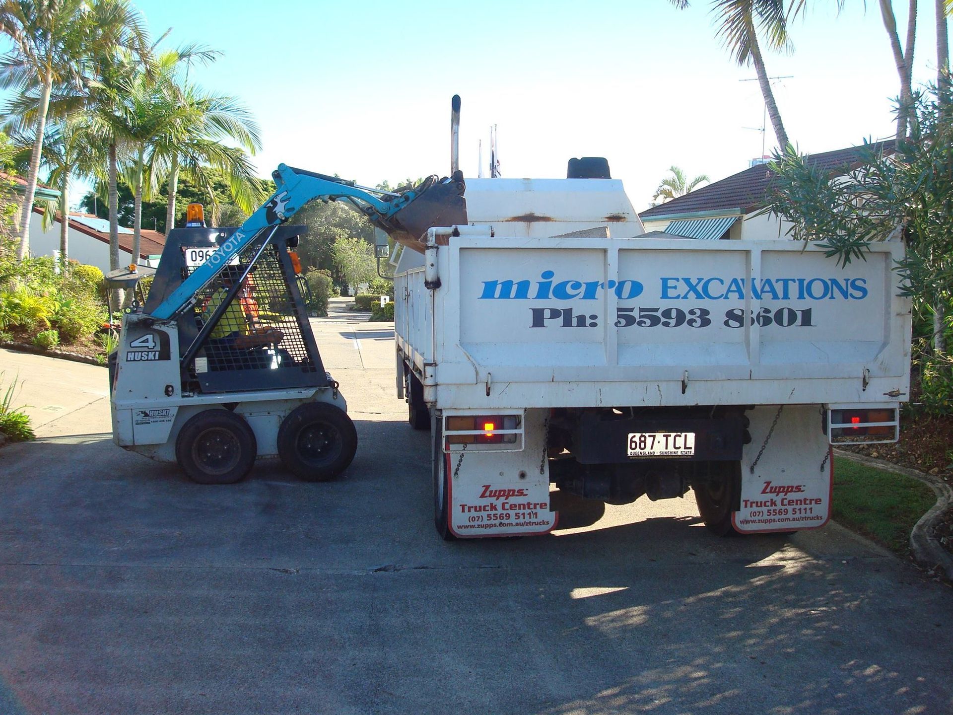 A small excavator loading dirt into the bed of a white Micro Excavations truck parked on a driveway.