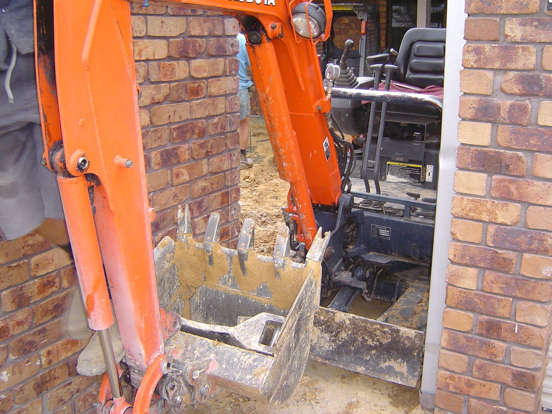 Orange excavator bucket digging a trench between brick walls.