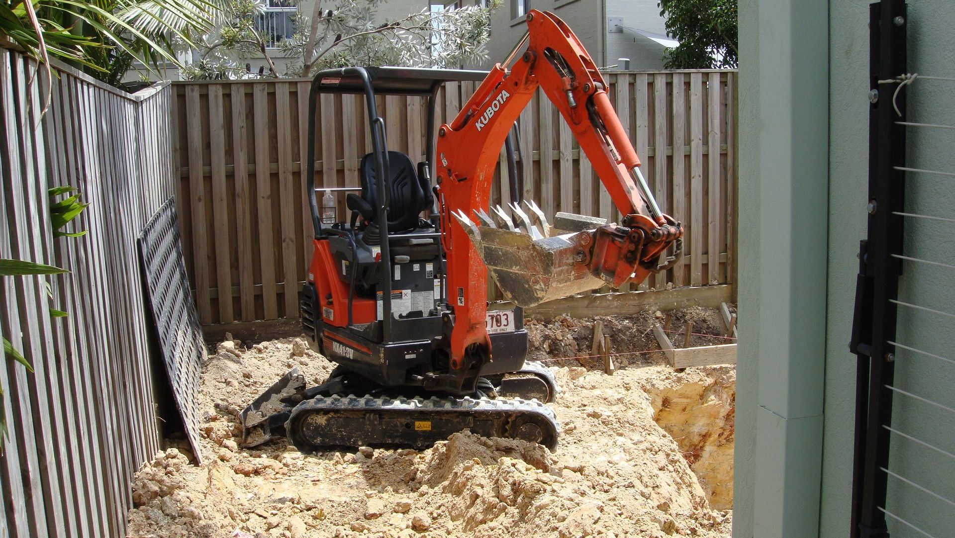 Orange Kubota mini excavator digging in a sand pit next to a wooden fence.