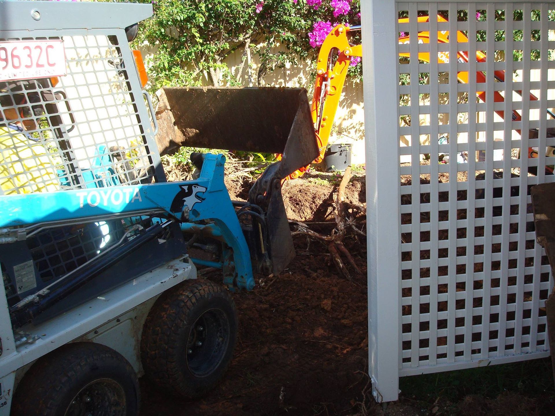 Blue Toyota skid steer operating in a yard, removing soil next to a white lattice fence.