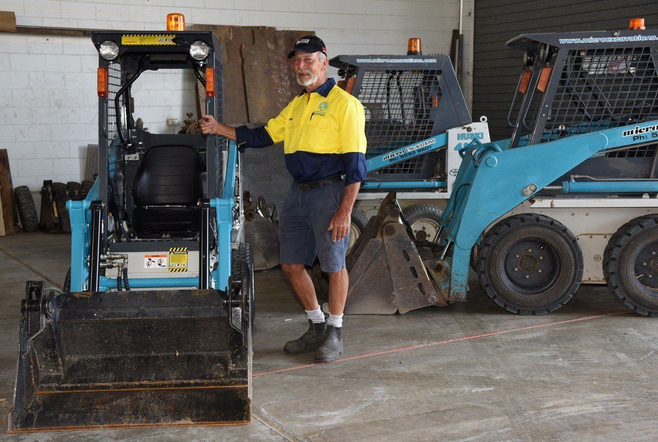 Man in work clothes poses next to a light blue skid steer loader in a garage.