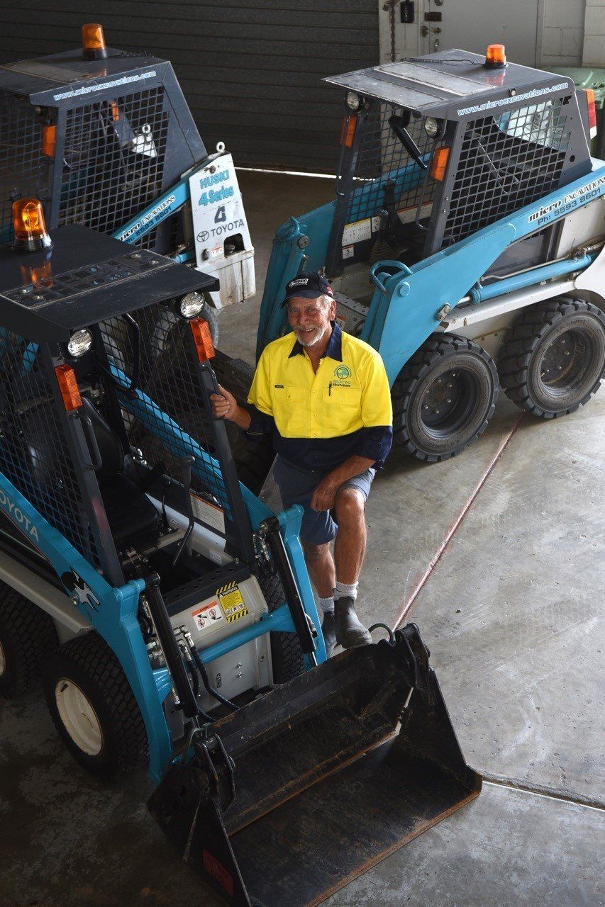 A man in yellow shirt sits between two light-blue skid-steer loaders in a garage-like setting.