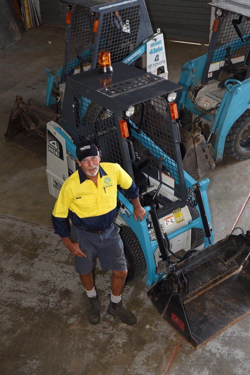Man standing in front of two skid steer loaders; he wears a yellow and blue shirt and shorts.