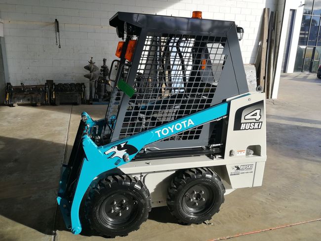 Toyota skid steer loader, gray and turquoise, with cage, on a concrete surface.