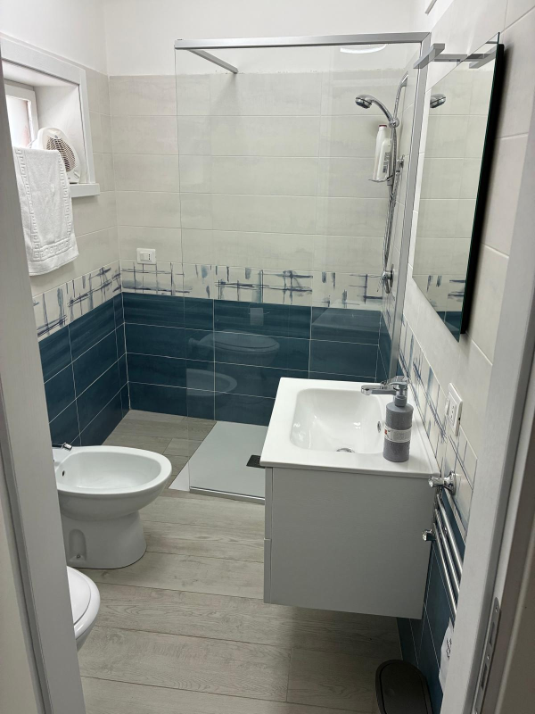 Piccolo bagno con piastrelle blu, WC, mobiletto sotto il lavabo e doccia sopra una vasca piastrellata.