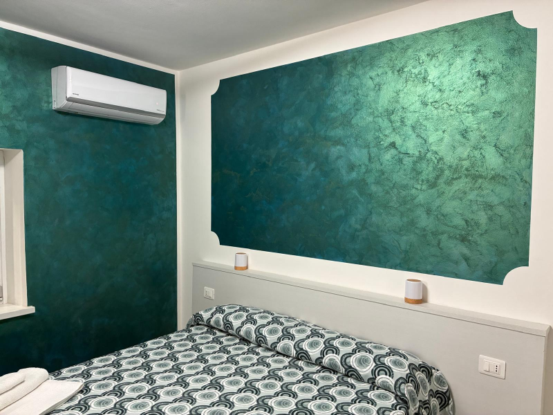 Letto ben rifatto con copriletto a fantasia color verde acqua e asciugamani bianchi arrotolati, sormontati da fiori rosa.