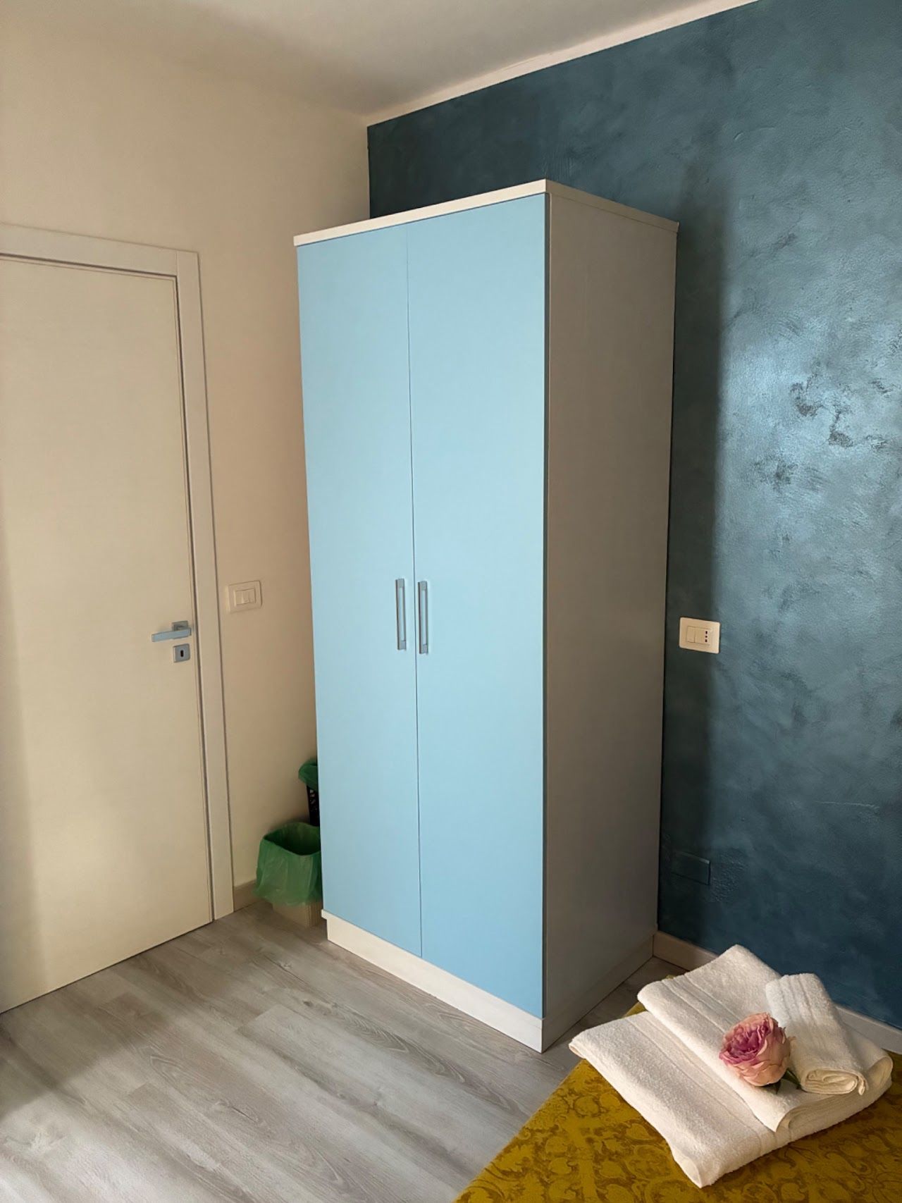Piccola camera da letto con armadio azzurro, porta bianca, parete color verde acqua e asciugamani piegati sul letto.