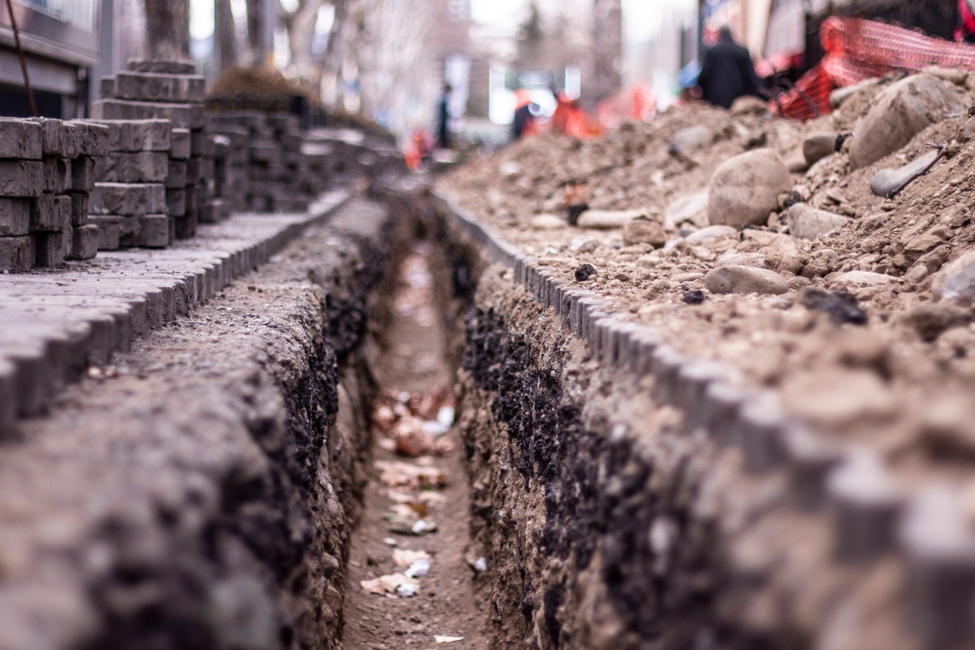Trench dug along sidewalk, with stacked pavers, dirt pile, and construction cones visible in the background.