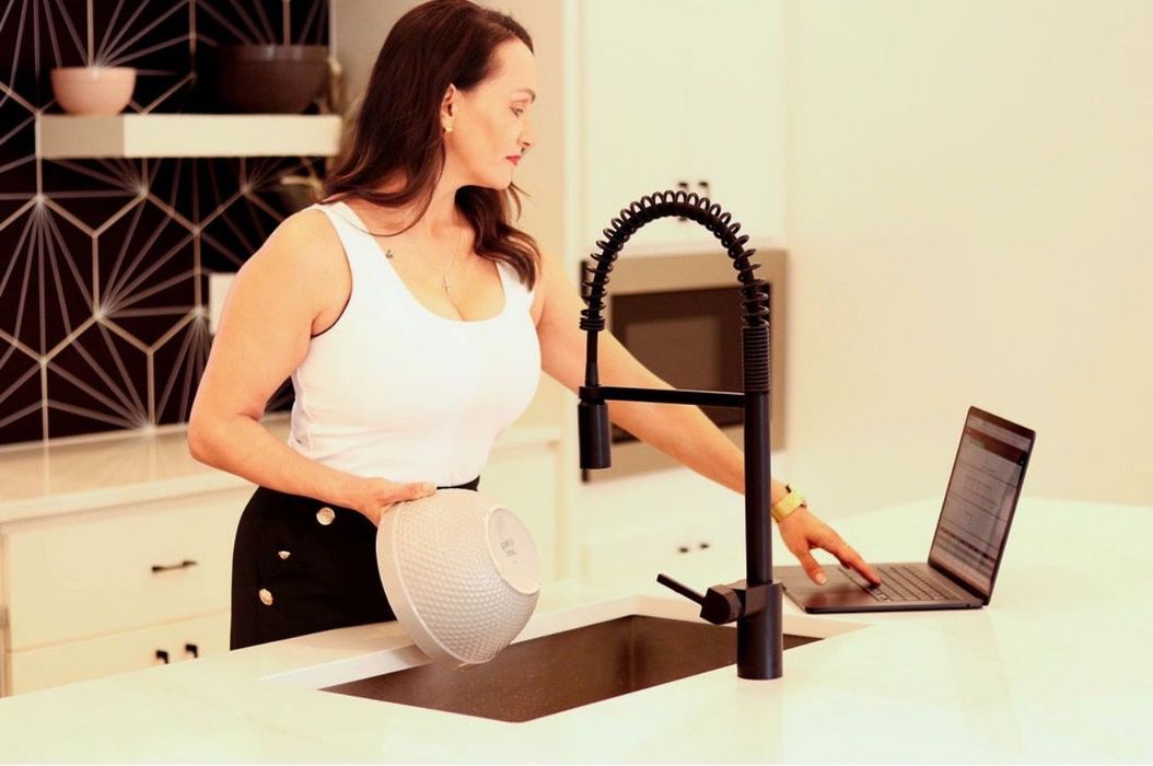 Woman washing a dish at a kitchen sink beside an open laptop