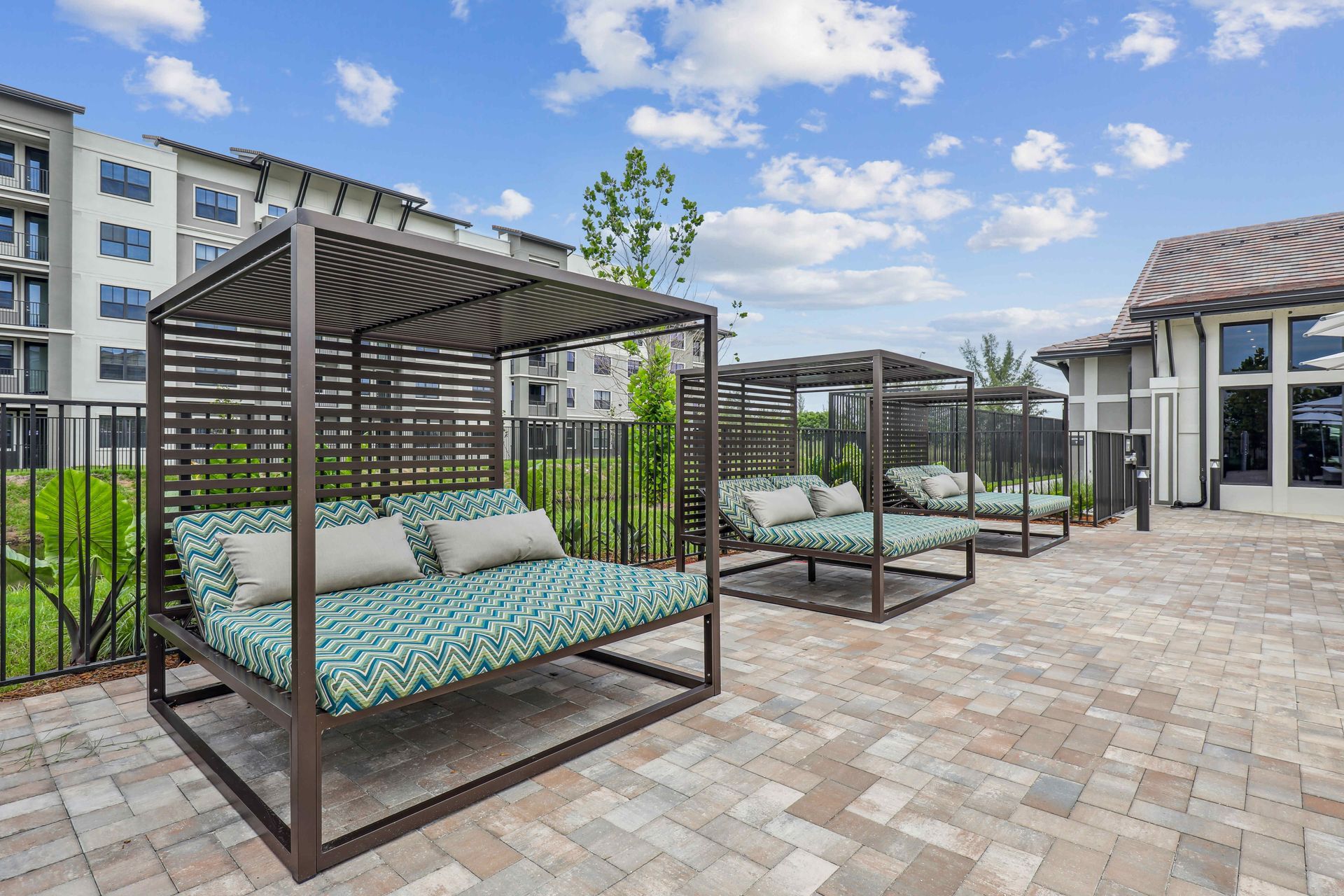 Outdoor lounge area with shaded daybeds on a brick patio, near a fence and buildings.