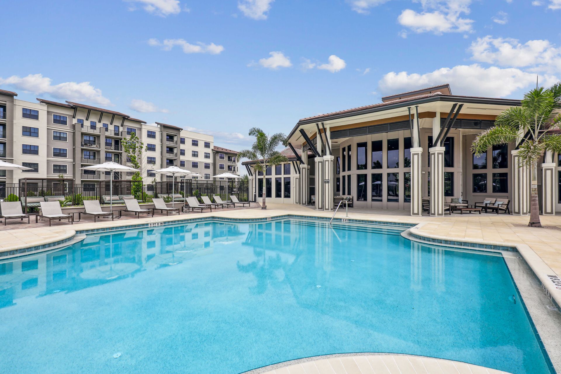 Swimming pool with lounge chairs, building, and palm trees under a blue sky.