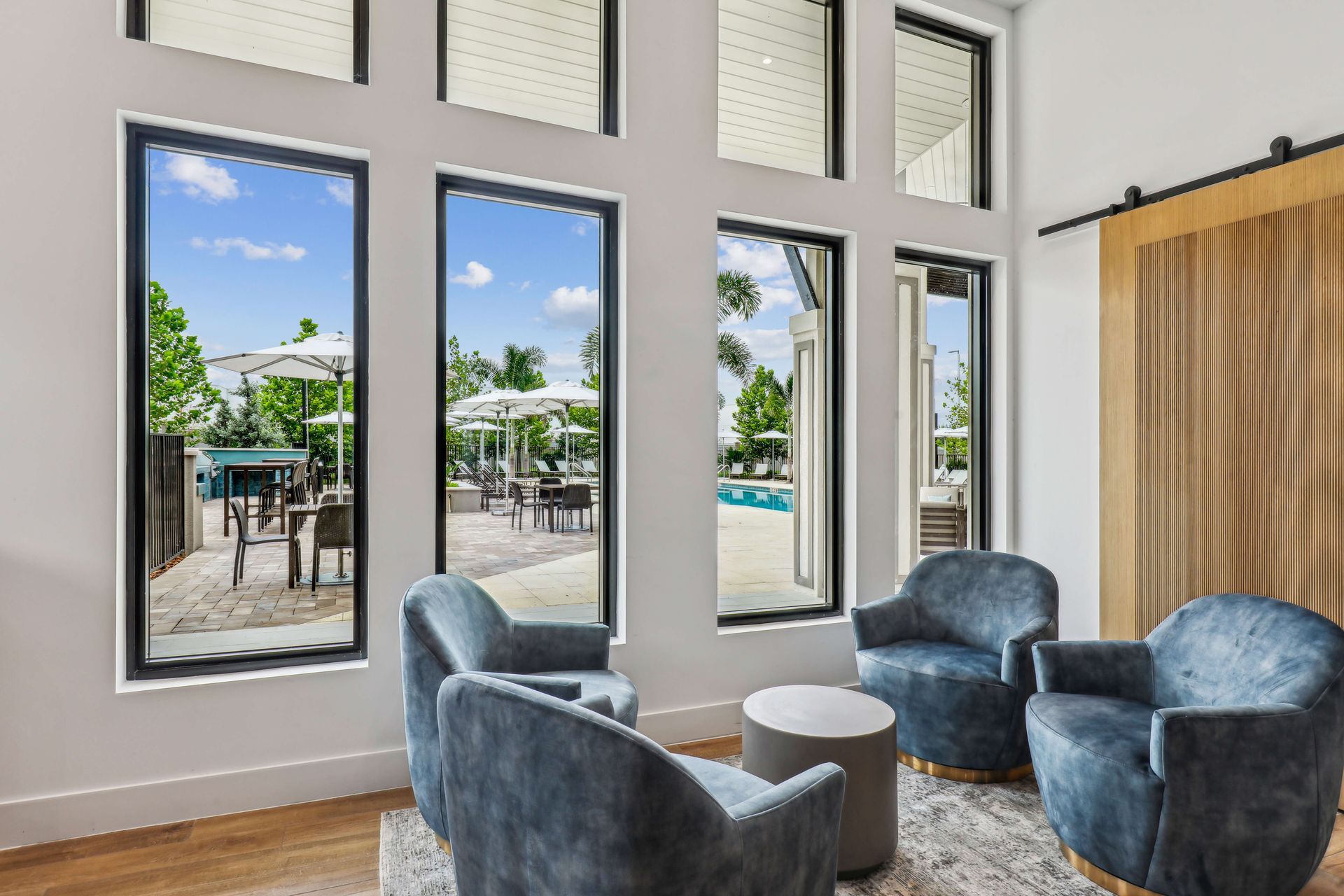 Living room with blue chairs, small table, and tall windows overlooking a patio with tables and a pool.