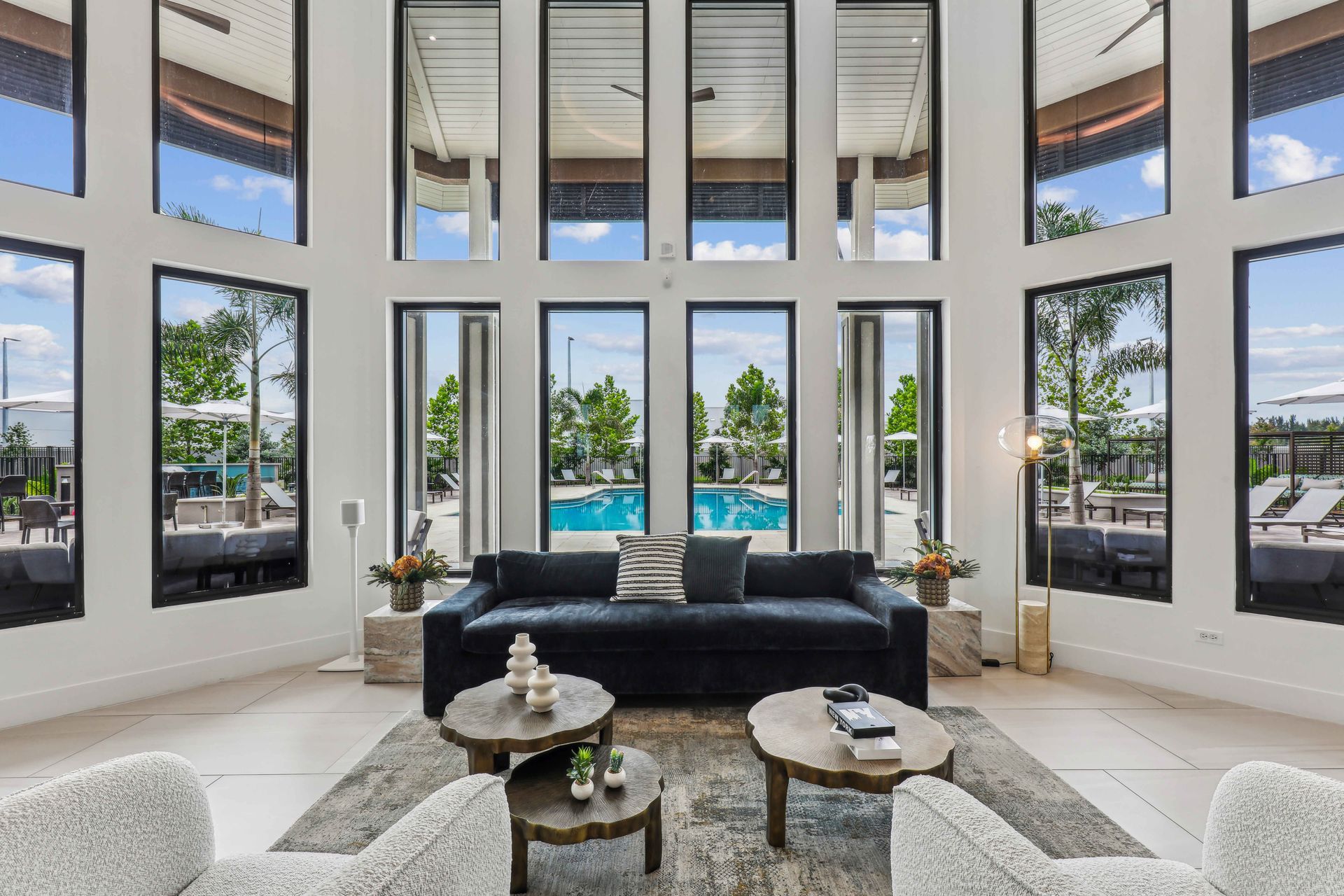 Spacious lounge with large windows overlooking a pool. Features a dark blue sofa, light chairs, and coffee tables.