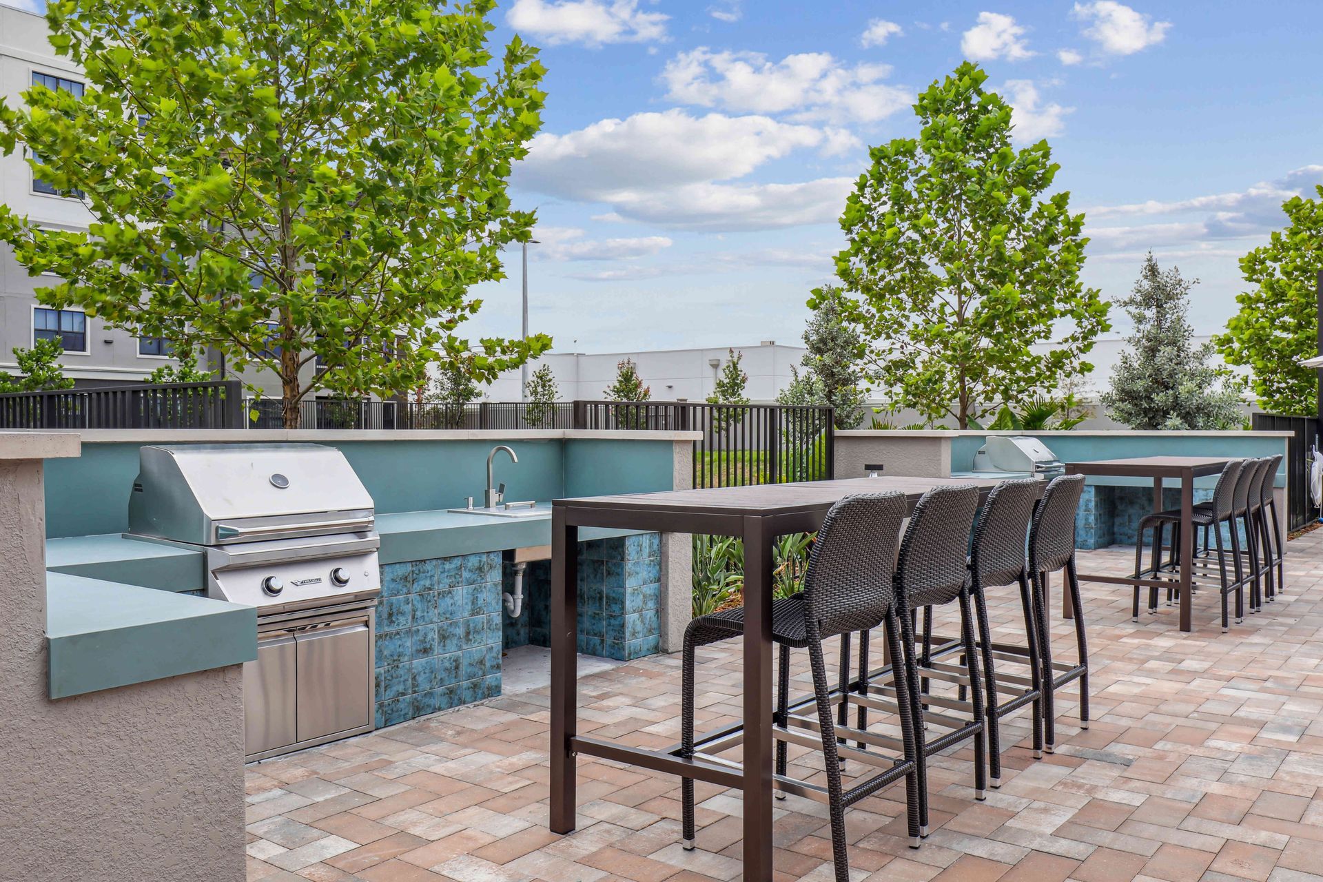 Outdoor grilling area with built-in grill, countertops, and high-top tables. Trees and blue tiled accents.