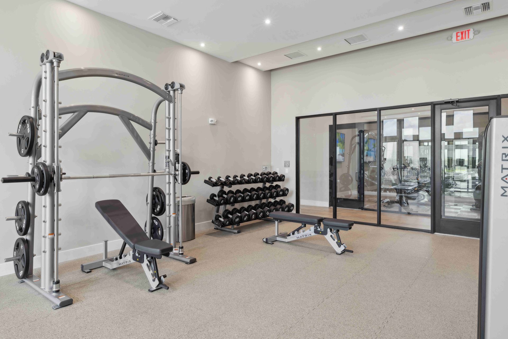 Gym with weight machines, dumbbells, and benches. Light grey walls, neutral floor.