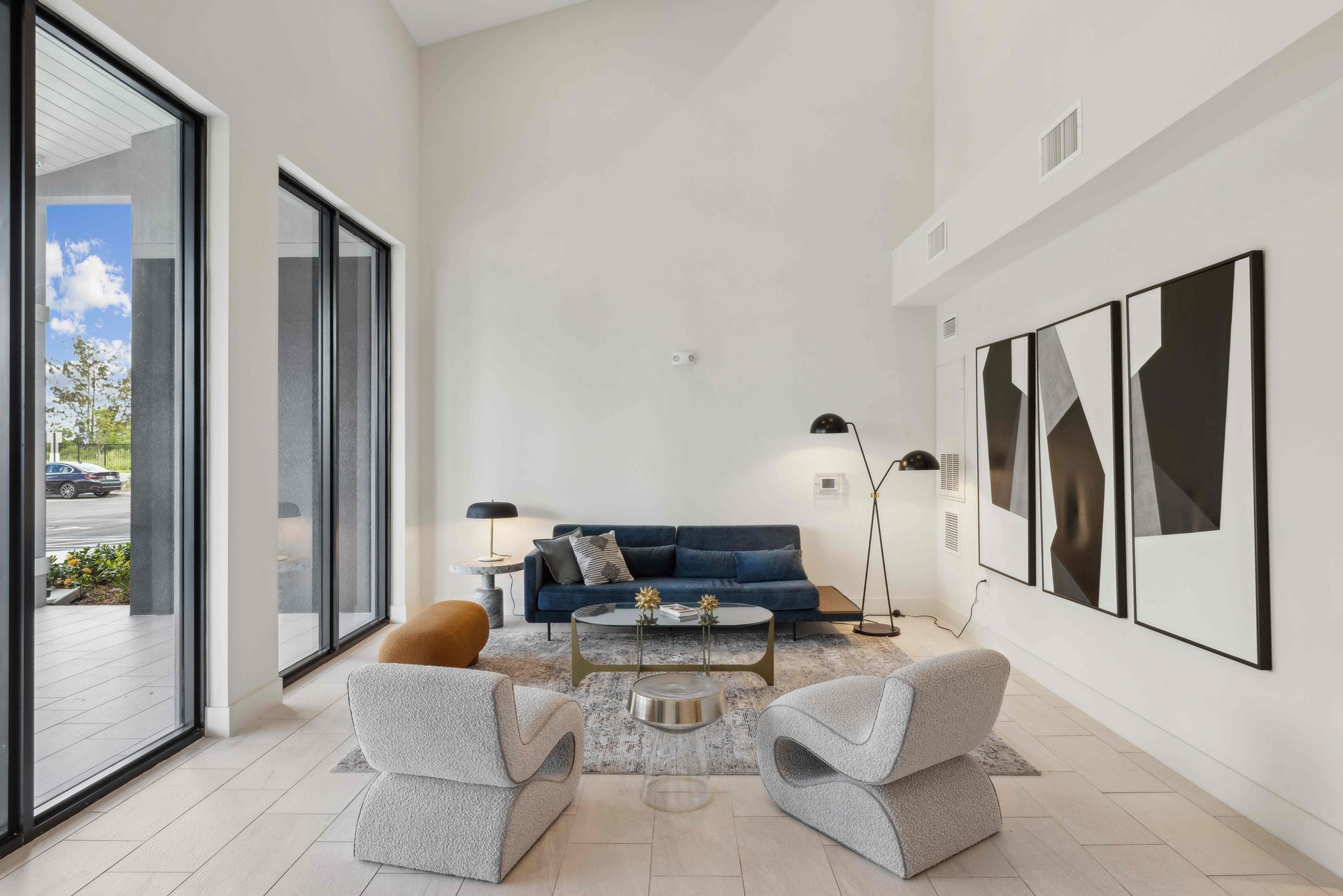 Modern living room with high ceiling, large windows, blue sofa, two patterned chairs, and abstract art.