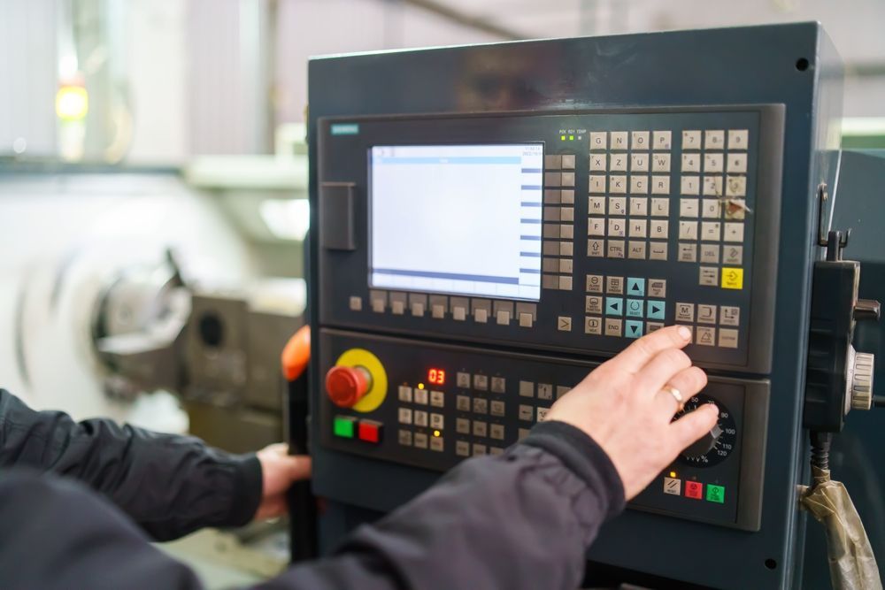 Hands operating a CNC machine control panel with buttons, screen, and emergency stop