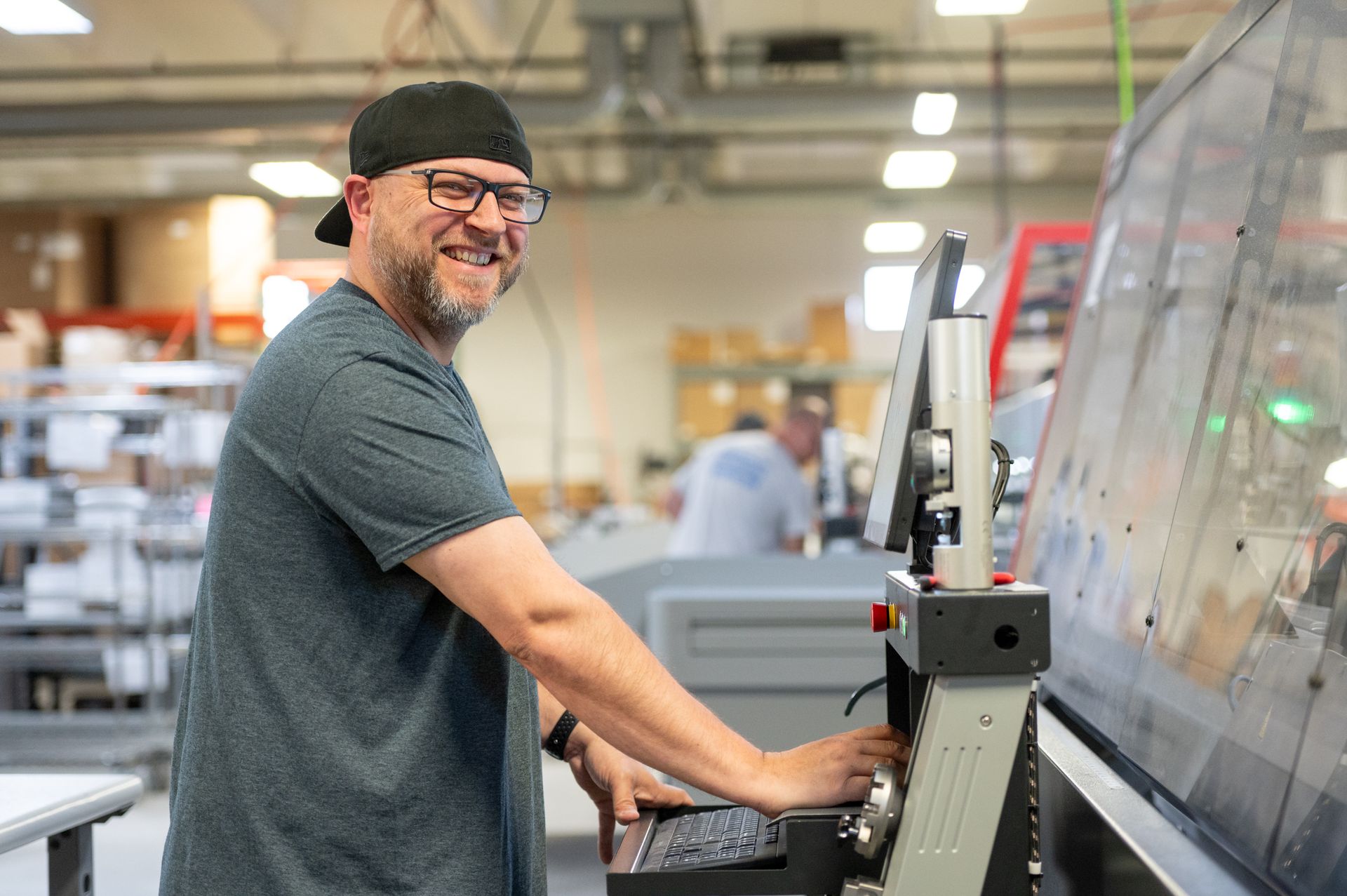 A male Kato Cable technician standing next to a wiring machine, smiling.