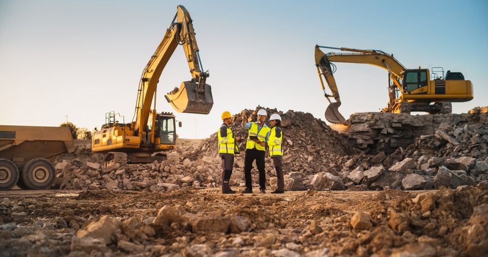 Three construction workers in vests and hardhats on a work site, with excavators and a dump truck.