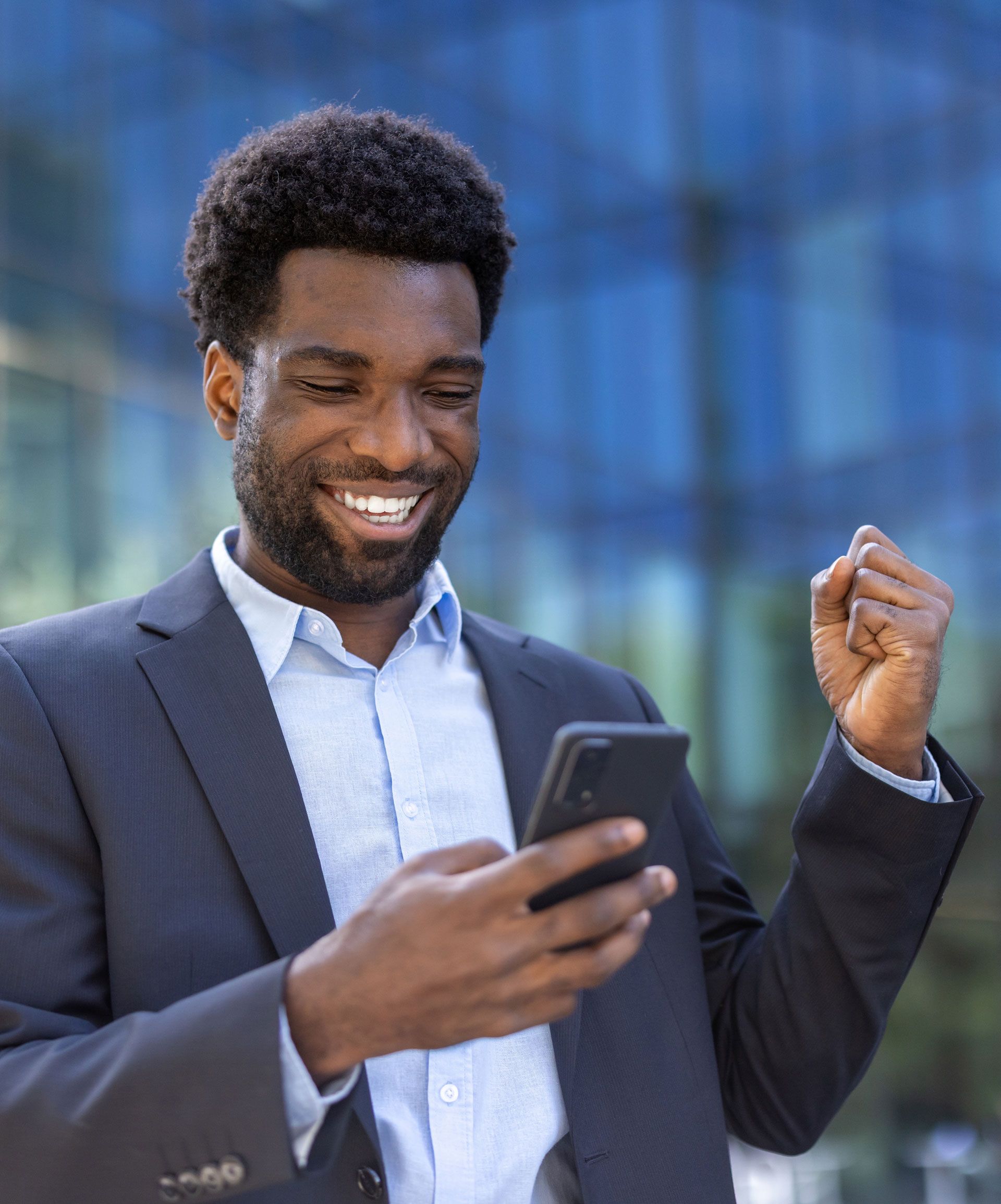 Man in suit, smiling while looking at a phone, fist raised in front of a building.