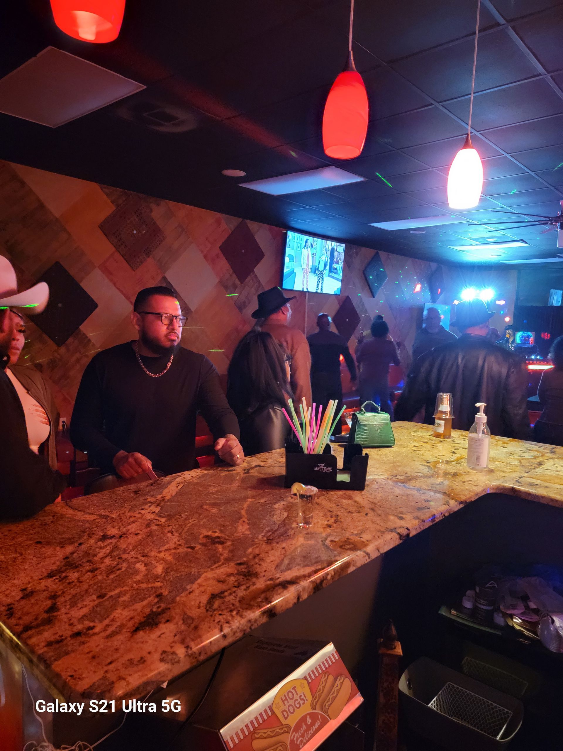 People at a bar with dim lighting, brown countertop, and red hanging lights.