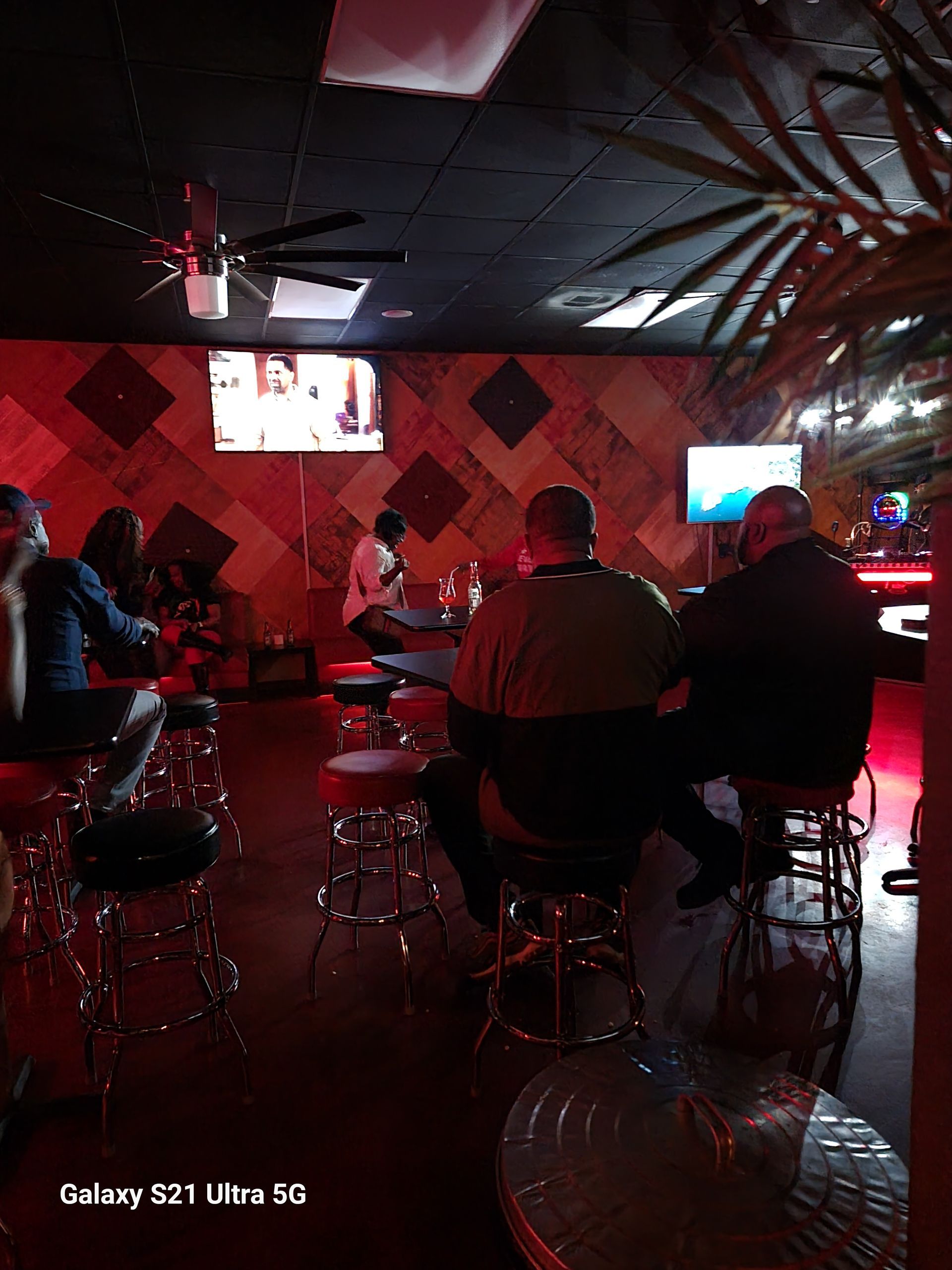 People seated at barstools inside a dimly lit bar with red lighting, watching TVs on the wall.