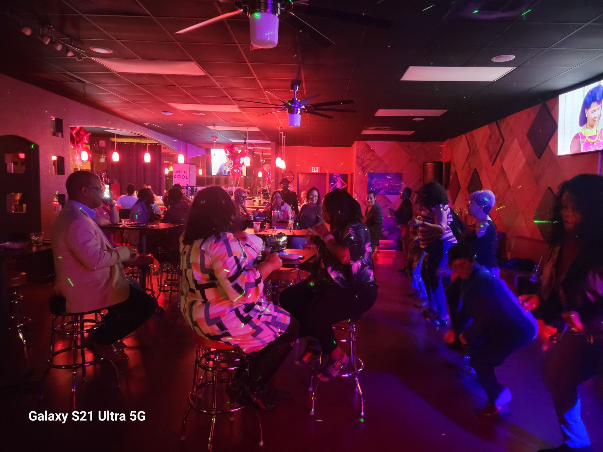 People socializing in a dimly lit bar with red and purple lighting. Some are seated, others dancing.