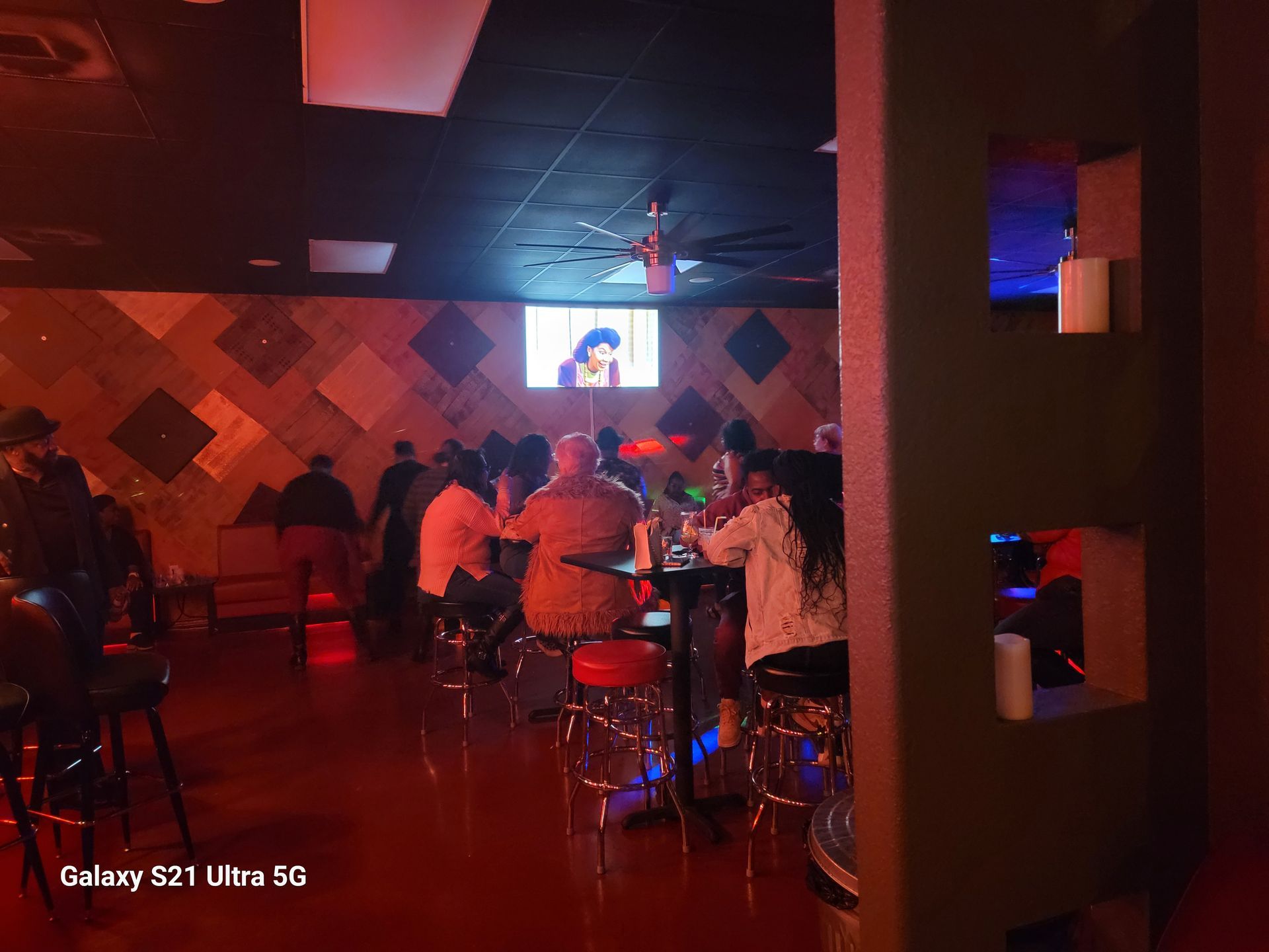 People in a dimly lit lounge, watching a screen. Red and dark hues dominate the scene, with visible bar stools and tables.