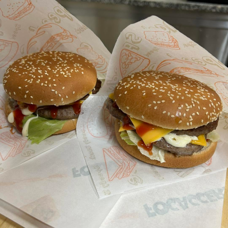 Due hamburger con semi di sesamo avvolti nella carta, uno con formaggio e pomodoro, su un vassoio.