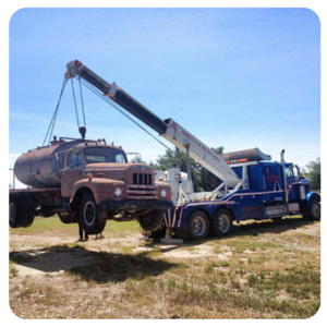 Tow truck lifting an old tan truck in a grassy field under a blue sky