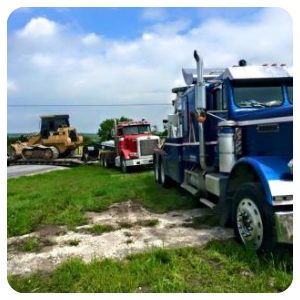 Blue semi-truck in foreground with red truck and bulldozer lined up on a grassy roadside under a cloudy sky
