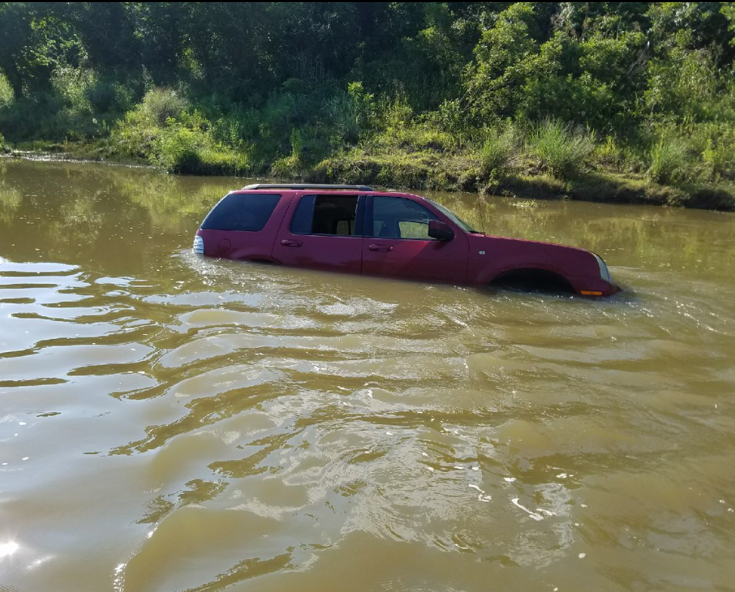 Magenta SUV partially submerged in muddy water near a wooded riverbank