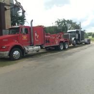 Red tow truck hauling a black semi-truck on a roadside under cloudy sky