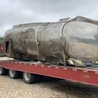 Large wrapped cylindrical object strapped to a red flatbed trailer in a gravel lot