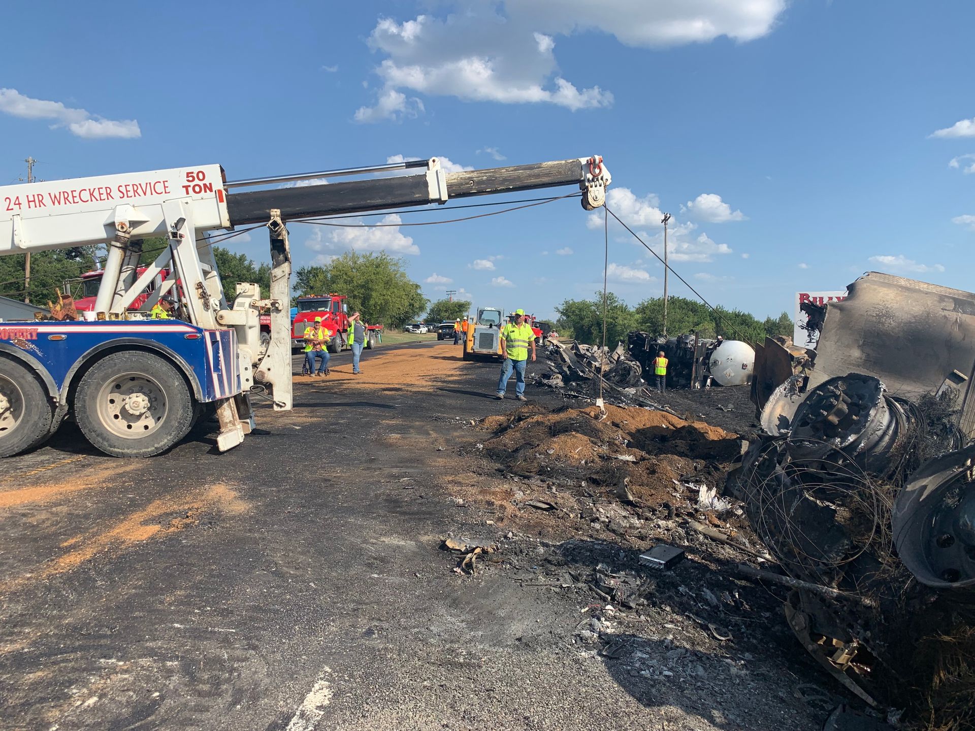 Tow truck at roadside crash scene with firefighters, smoke, and a burned-out vehicle