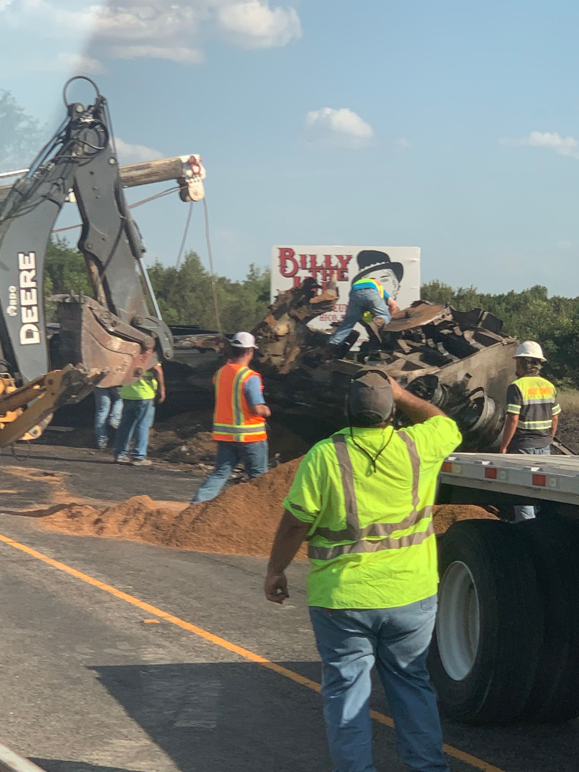 Roadwork crew clearing spilled dirt on highway beside a black excavator and truck under a blue sky