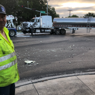 Highway crash scene with a white tanker truck and debris, viewed from a sidewalk with a worker in a safety vest.
