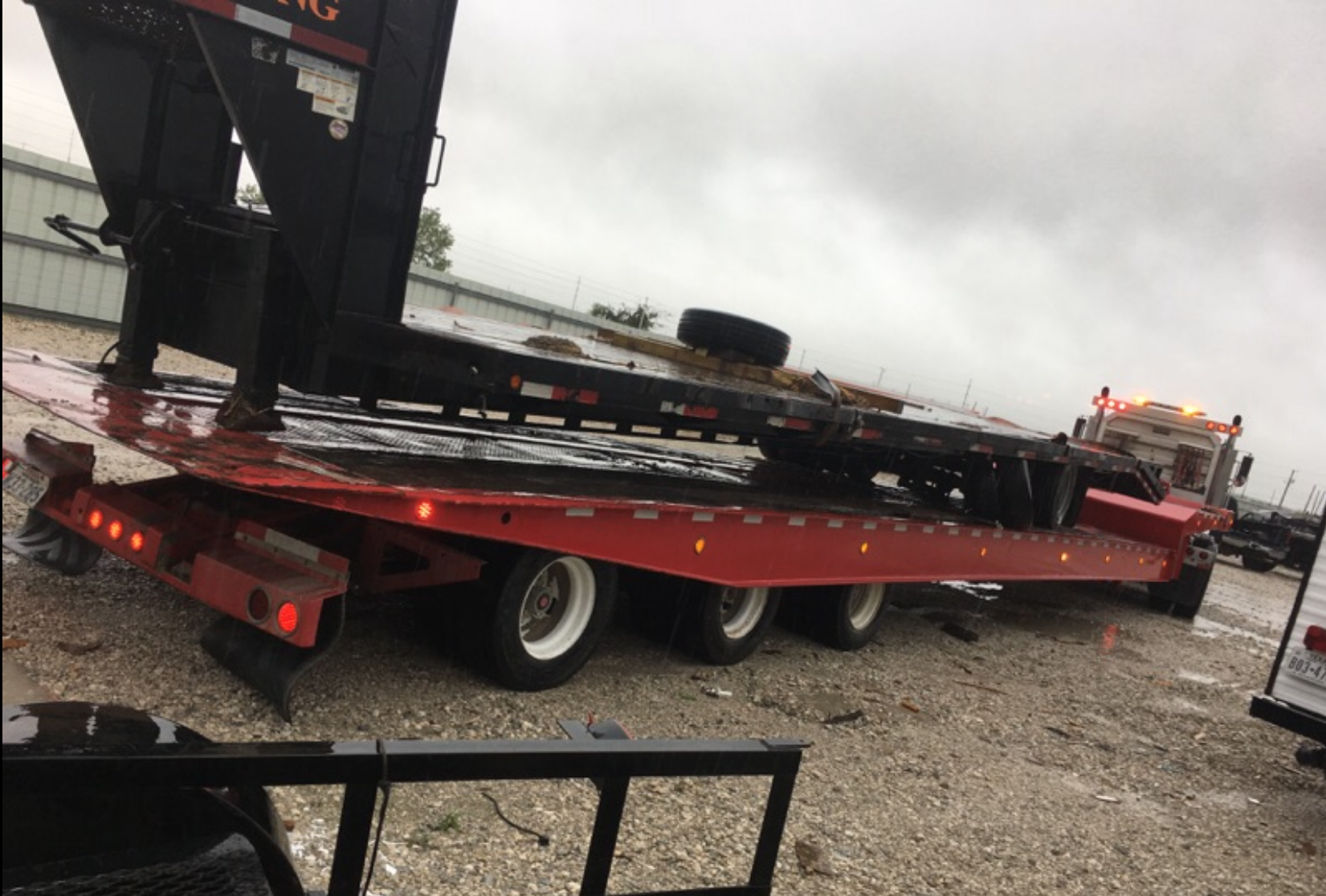 Red flatbed tow trailer loaded with equipment on a gravel lot in rainy weather