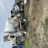 Overturned white cement mixer truck in a ditch beside a blue semi-truck on a grassy roadside.