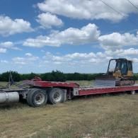 Red flatbed semi-trailer parked in a field under a cloudy sky, with a bulldozer on the trailer.