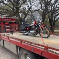 Motorcycle strapped on a red flatbed tow truck outside a red building under leafless trees
