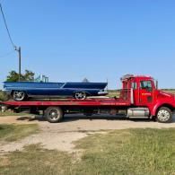 Red flatbed tow truck carrying a blue classic car on a sunny roadside.